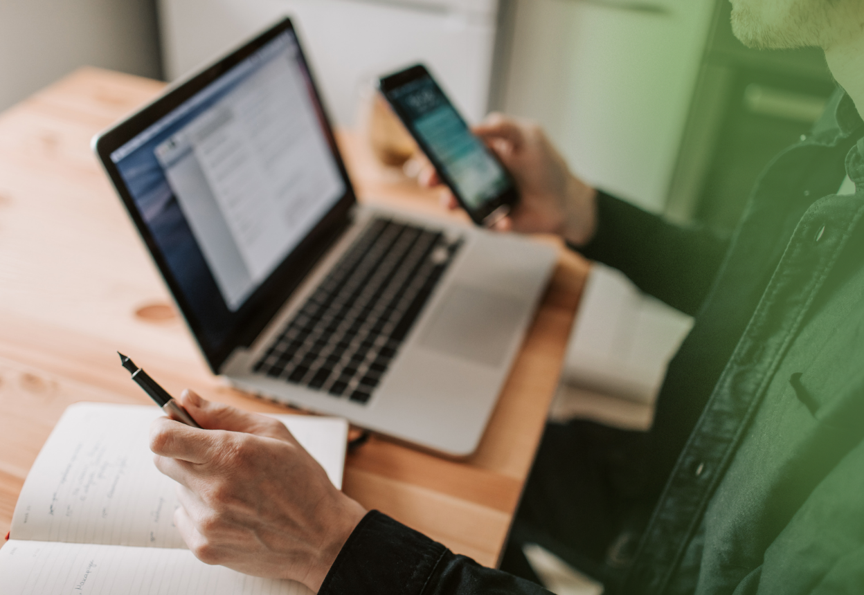 A man is sitting at a table with a computer and a notebook on it; he is holding a phone in one hand and a pen in the other
