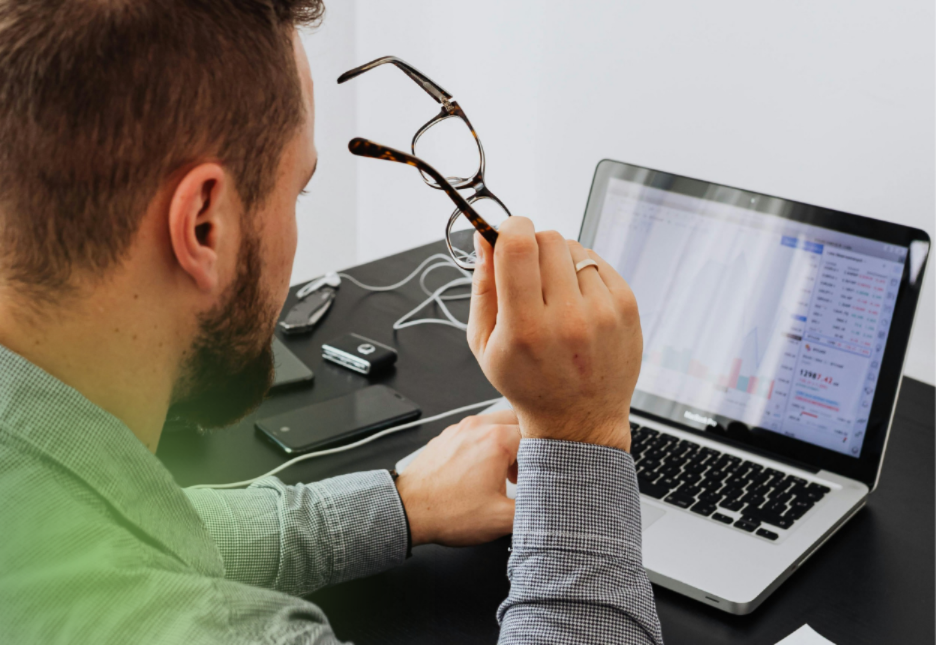 A man sitting at a table, holding his glasses in his hands, is looking at the computer screen